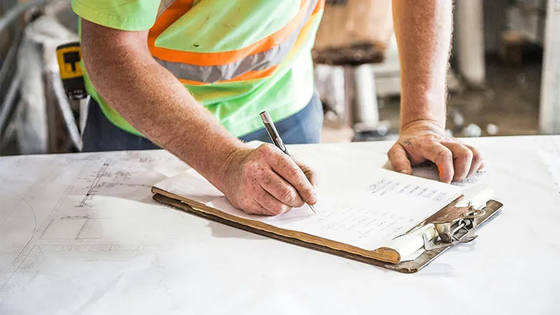 A construction worker is writing with a pen and signing a document on a clipboard.