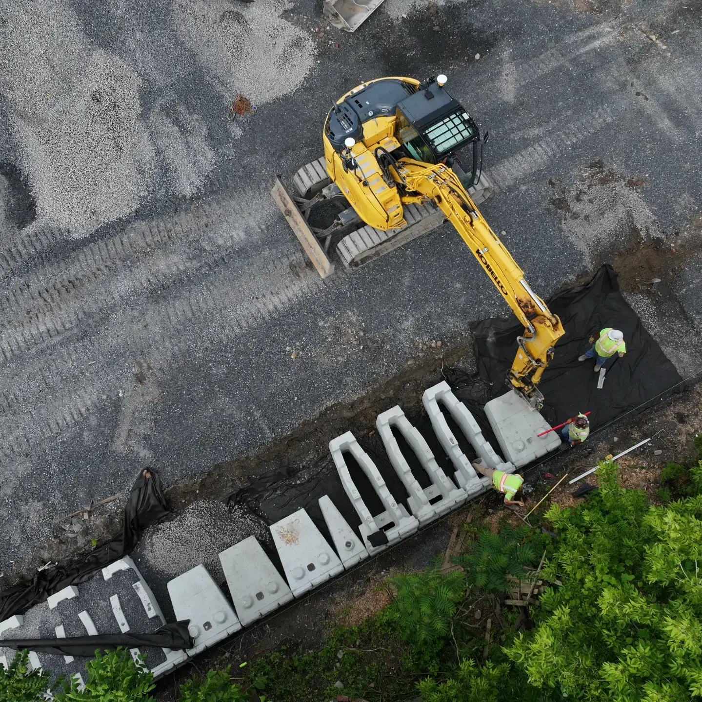 An aerial view of a construction site with an excavator and workers amidst dirt mounds.